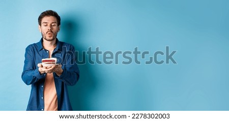 Celebration and holiday concept. Handsome young man blowing candle on birthday cake and making wish, standing on blue background.