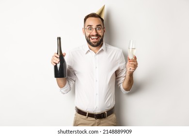 Celebration and holidays. Excited man enjoying birthday party, wearing b-day hat and drinking champagne, standing over white background