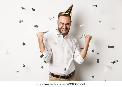 Celebration and holidays. Happy man dancing on birthday party with confetti, wearing b-day hat and rejoicing, standing over white background
