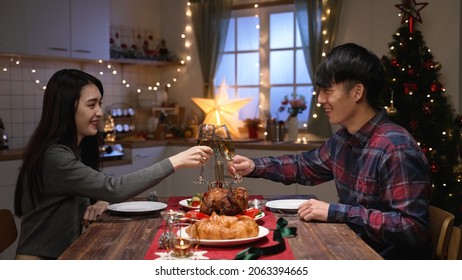 Celebration People And Holidays Concept. Smiling Young Asian Couple Clinking Glasses Of Alcoholic Wine At Home Over Christmas Tree Background. Sweet Lovers Enjoy Xmas Dinner Indoors