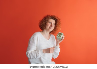 Celebration. Portrait Of Happy Looking Red-haired Woman Holding Small Gift Box Isolated Over Red Studio Background. Birthday Present. Concept Of Facial Expression, Emotions, Feelings, Ads