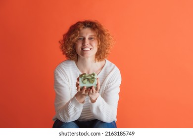 Celebration. Portrait Of Happy Looking Red-haired Woman Holding Small Gift Box Isolated Over Red Studio Background. Birthday Present. Concept Of Facial Expression, Emotions, Feelings, Ads