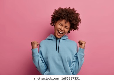 Celebration, success and triumph concept. Carefree joyful Afro American woman makes fist bump, celebrates victory, yells hooray as win prize, dressed in blue casual hoodie, isolated on pink background