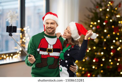 Celebration, Technology And Winter Holidays Concept - Happy Couple In Santa Hats Taking Picture By Smartphone On Selfie Stick At Ugly Sweater Party Over Christmas Tree At Home Background