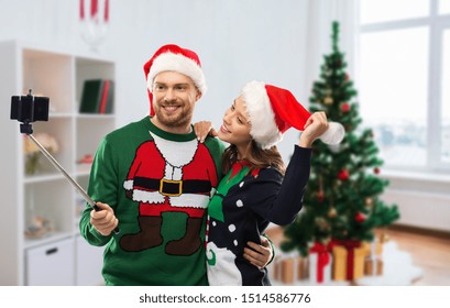 Celebration, Technology And Winter Holidays Concept - Happy Couple In Santa Hats Taking Picture By Smartphone On Selfie Stick At Ugly Sweater Party Over Christmas Tree At Home Background