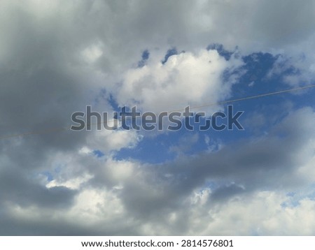 Celestial contrast of dark cumulus clouds and blue sky with minimal powerline.
A captivating, low-angle shot of a dynamic sky featuring a mix of heavy grey storm clouds and bright white patches against a deep blue background. A single, thin, almost unseen powerline cuts diagonally across the middle, adding a subtle touch of human presence to the immense natural atmosphere.