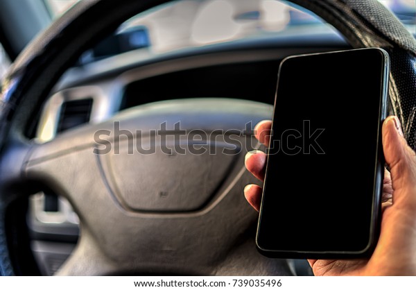Cell phone with blank screen in hand on car\
steering wheel background. Black screen of phone for your\
inscription or pictures.