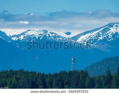 Cell tower rises above tree line of temperate rainforest near a building (bottom left) along Auke Bay, with view of snow-capped mountains and low clouds, on an afternoon in mid June, southeast Alaska