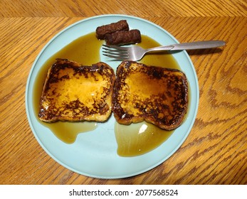 A cellphone image of a home made breakfast made with French toast covered in syrup with two link sausage salted and served with a fork on a blue plate on a wooden table top