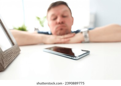 Cellphone lying on table with pensive man in background waiting for call or out of network coverage closeup