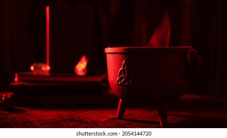 Celtic Witch Ritual Cauldron With Spell Books And Crystals Over Black Table With Magic Wand In Eerie Red Light