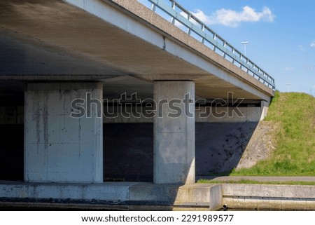 Cement concrete pillar structure of urban bridge crossing over small river, canal or ditch with green grass meadow, Underneath of classic style bridge under blue sky and white clouds, Netherlands.