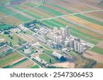 Cement plant in Austria view from above. Concrete plant, surrounded by agricultural fields. Facility includes multiple tall silos, various buildings, and infrastructure such as roads and rail tracks