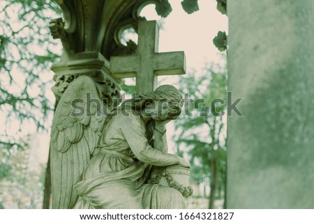 cementery angel looking down in green tone, gray stone cross, tombestone, sadness concept