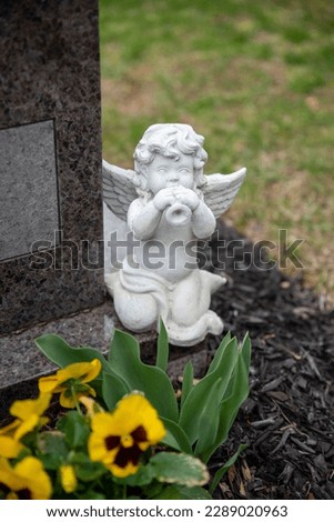 A cemetery headstone with a white cherub angel statue with wings. Yellow flowers at the base of the head stone, green grass background. No people, with copy space.