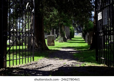 Cemetery with iron gate entrance, sun and shadow pathway, tombstones, and trees. - Powered by Shutterstock