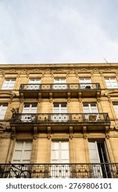 Centered low-angle portrait shot looking up at three consecutive floors of a classic Haussmann-inspired building facade in Bordeaux, France. Each level features matching black wrought iron balconies and tall French windows framed by ornate stone moldings, creating a strong sense of repetition and verticality. A narrow strip of clear blue sky crowns the top of the composition, emphasizing the imposing height and elegance of this limestone facade.
