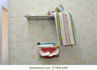 A centered shot showcasing a floral patterned soap dish holding two bars of white soap, a tube of toothpaste, and a green toothbrush attached to a tiled wall next to a striped hanging towel.