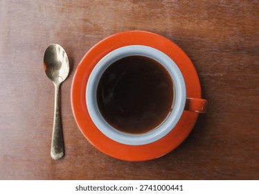 A centered top-down shot of a red cup filled with black coffee, accompanied by a spoon on a rustic table. The image relies on natural lighting to convey a sincere, everyday lifestyle moment.