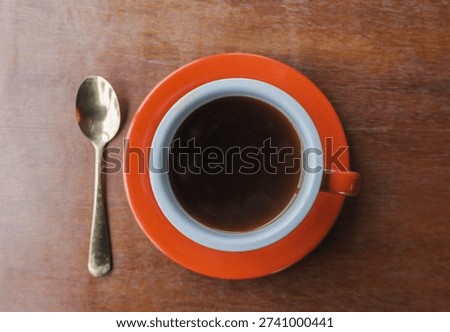 A centered top-down shot of a red cup filled with black coffee, accompanied by a spoon on a rustic table. The image relies on natural lighting to convey a sincere, everyday lifestyle moment.