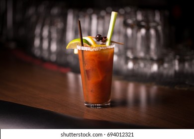 Centered Traditional Vodka Tomato Juice Bloody Mary Mix Cocktail In Salted Rim Tumbler Garnished By Lemon With Olive And Celery Stalk On Retro Diner Bar With Blurry Stacked Glass Restaurant Background