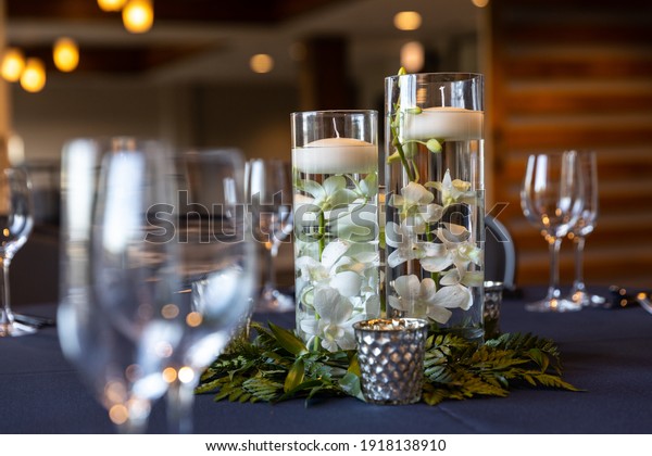 A centerpiece at a wedding with wine glasses on\
the table, flowers and floating candles in pillar vases, and\
blurred out lights in the\
distance.