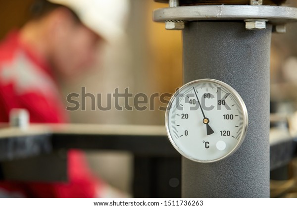 Central heating network. Thermometer on the hot\
water supply pipe to the heating system. Technician wearing red\
jacket with white helmet blurred in background. Start of the\
heating season.