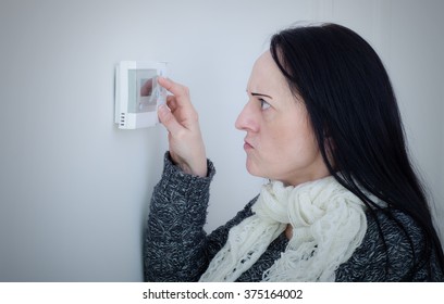 Central Heating Problems. Cold And Unhappy Woman Struggling To Turn Heating Temperature Up On Wall Thermostat.