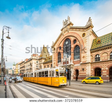 Central Market Hall in Budapest city, Hungary, Europe. Pedestrian crossing and yellow tram in foreground, old building and blue sky in background.