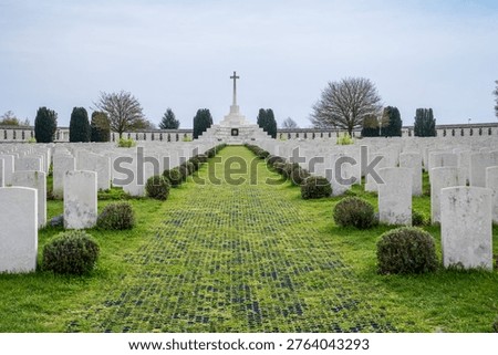 A central perspective of Tyne Cot Cemetery in Belgium. Symmetrical rows of white headstones on green grass lead to the Cross of Sacrifice. A solemn Commonwealth site dedicated to the fallen of the Great War, a UNESCO World Heritage site.