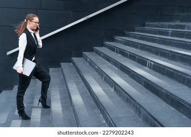 CEO woman talking on smartphone, or secretary goes to work, climb stairs. A woman in a black suit and white shirt holds a laptop in her hands. Caucasian business successful woman - Powered by Shutterstock