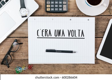 C'era una volta, Italian text for Once Upon a Time, on note pad at office desk with electronic devices, computer and paper, wood table from above, concept image for blog title or header image.