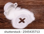 A ceramic bowl of sugar spilled on a wooden table with an "X" symbol drawn in the sugar and a wooden spoon filled with sugar placed nearby