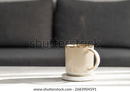 Ceramic cup of coffee on light table with morning shadows, in front of a gray sofa. Concept of cozy and minimal home decor scene. Front view, copy space.