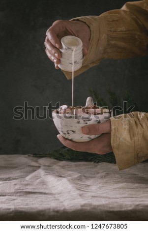 Ceramic cup of hot chocolate with marshmallow s'mores in female hands. Pouring cream from jug. Grey linen table cloth. Winter drink. Dark atmosphere.