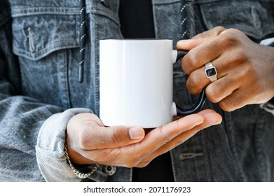 Ceramic mug mockup. Close up african-american in a denim jacket holding white cup with black handle of coffee, copy space. Front view, space for branding imprint