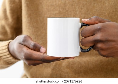 Ceramic mug mockup. Close up african-american hands holding white cup with black handle of coffee, copy space. Front view, space for branding imprint