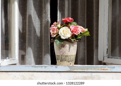 Ceramic Partially Broken Flower Vase With Plastic Roses In Various Colors On Top Of Window Seal With Open Windows And Curtain In Background On Warm Sunny Day