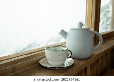 Ceramic pot and cup sit elegantly on wooden ledge with snowy mountains outside.