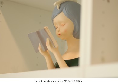 Ceramic Statue Of A Woman Reading A Book Placed On A White Shelf. Close Up Plastic Statue Of A Lady With Short Hair.