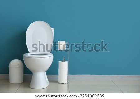 Ceramic toilet bowl, bin and paper rolls near blue wall