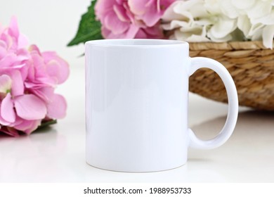 Ceramic white coffee mug with pink and white flowers in the wicker basket. Blank 11 oz mug mockup for design.