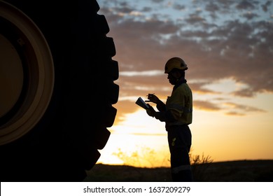 Certified  Motor Vehicle Mechanic Mining Haul Truck Driver Standing Holding Inspection Pre Operation Checklist Book Inspecting Taking Pictures Of Defect, Damage Of Wheel Tyre Sunset Background 