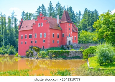 Cervena Lhota Castle gracefully rises from a rocky island, encircled by a serene lake. Lush greenery frames the vibrant structure under a clear blue sky. - Powered by Shutterstock