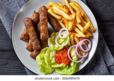 cevapcici, grilled balkan sausages with potato fries, red onion rings, ajvar and fresh lettuce leaves on a plate on a black wooden table, flat lay, close-up