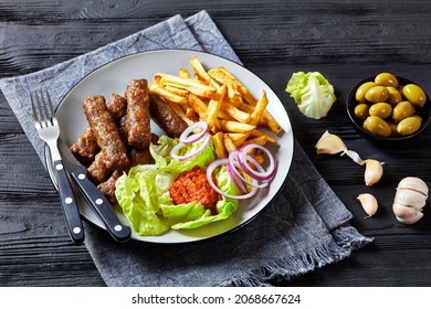 cevapcici, grilled balkan sausages with potato fries, red onion rings, ajvar and fresh lettuce leaves on a plate on a black wooden table with green olives, close-up