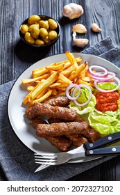 cevapcici, grilled balkan sausages with potato fries, red onion rings, ajvar and fresh lettuce leaves on a plate on a black wooden table with green olives, vertical view, close-up