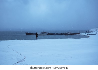 A chain of footprints in the newly fallen snow. Snowfall in the evening twilight. A lonely figure of a man looks at the blue water of Lake Baikal and the port of abandoned ships.  - Powered by Shutterstock