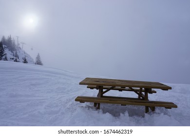 Chair Lift And Sun Seeing Through The Thick Fog By A Wood Picnic Table At The French Ski Resort Of L'Alpe D'Huez                        