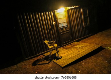 Chair On A Cabin Porch At Night: A Light Over A Cabin Door Illuminates An Empty Chair. Taken In Lexington, Michigan, USA.